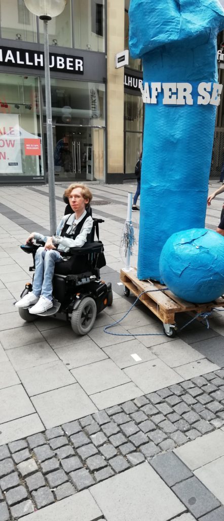 Ein Rollstuhlfahrer steht neben einem 3 Meter hohen Plastik-Penis, der Penis trägt die Aufschrift "Safer Sex"