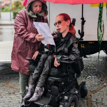 Auf dem Slutwalk stehen Slavica und ich am Gärtnerplatz in München vor dem Wagen des Slutwalks. Es regnet stark. Sie trägt eine rote Regenjacke mit Kapuze und blickt konzentriert auf die Rede, die sie für mich in ihrer rechten Hand hält, während ich spreche. In ihrer linken Hand hält sie mein Mikrofon. Ich trage eine schwarze Lederjacke, eine schwarze Lederhose und Stiefel mit einem Schlangenhautmuster. Meine roten Haare sind wetterbedingt zu einem Zopf gebunden. Ein roter Regenschirm, der mit einer Halterung an meinem Rollstuhl befestigt ist, schützt mit zumindest etwas vor dem Regen.