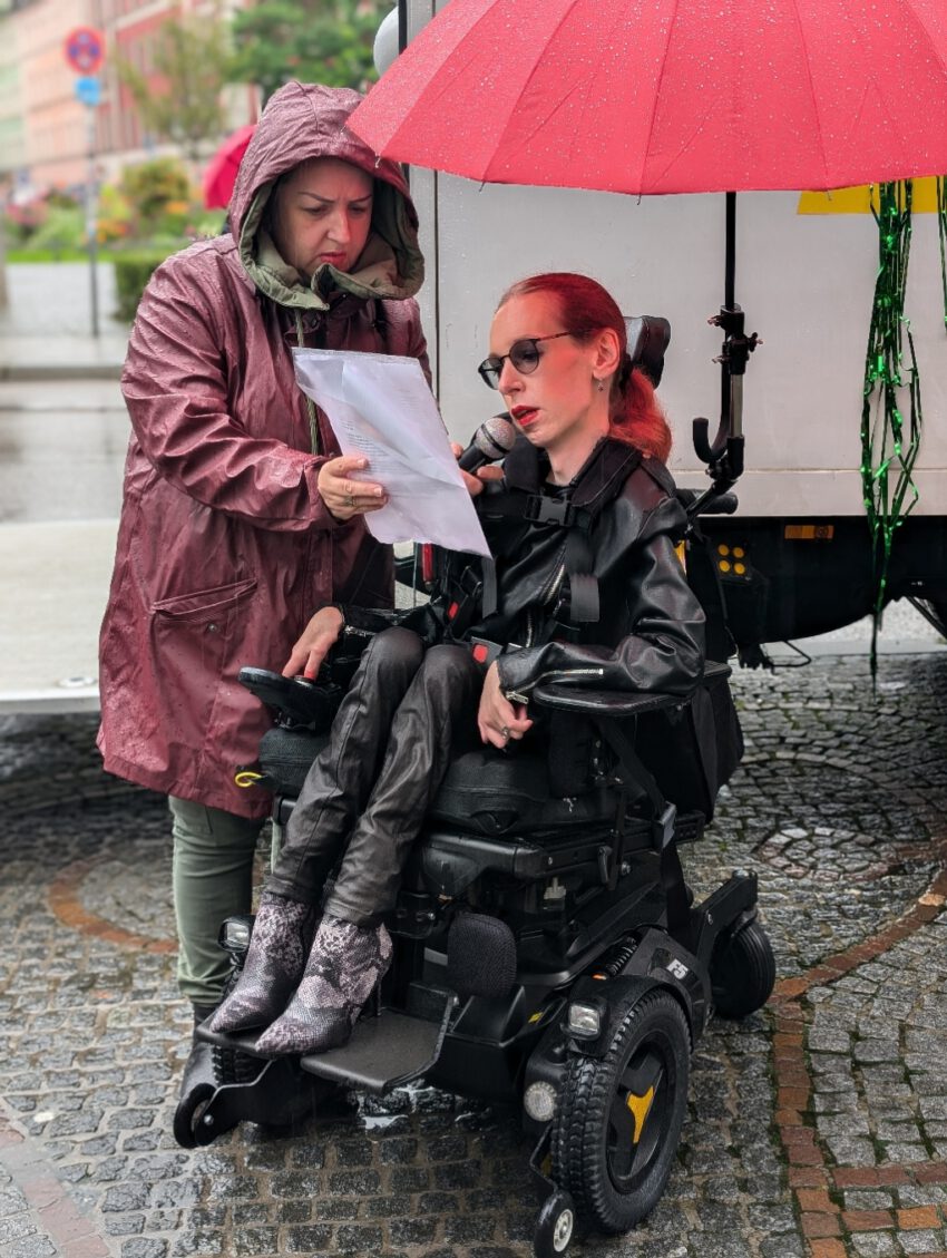 Auf dem Slutwalk stehen Slavica und ich am Gärtnerplatz in München vor dem Wagen des Slutwalks. Es regnet stark. Sie trägt eine rote Regenjacke mit Kapuze und blickt konzentriert auf die Rede, die sie für mich in ihrer rechten Hand hält, während ich spreche. In ihrer linken Hand hält sie mein Mikrofon. Ich trage eine schwarze Lederjacke, eine schwarze Lederhose und Stiefel mit einem Schlangenhautmuster. Meine roten Haare sind wetterbedingt zu einem Zopf gebunden. Ein roter Regenschirm, der mit einer Halterung an meinem Rollstuhl befestigt ist, schützt mit zumindest etwas vor dem Regen.