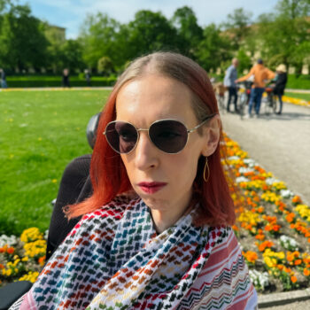 Ich stehe vor einem Blumenbeet, eingewickelt in eine bunte Kufiya. Mein Blick geht nachdenklich in die Ferne. Ich trage meine Haare offen und goldenen Schmuck. Mein Ausdruck ist äußerst nachdenklich und ernst.