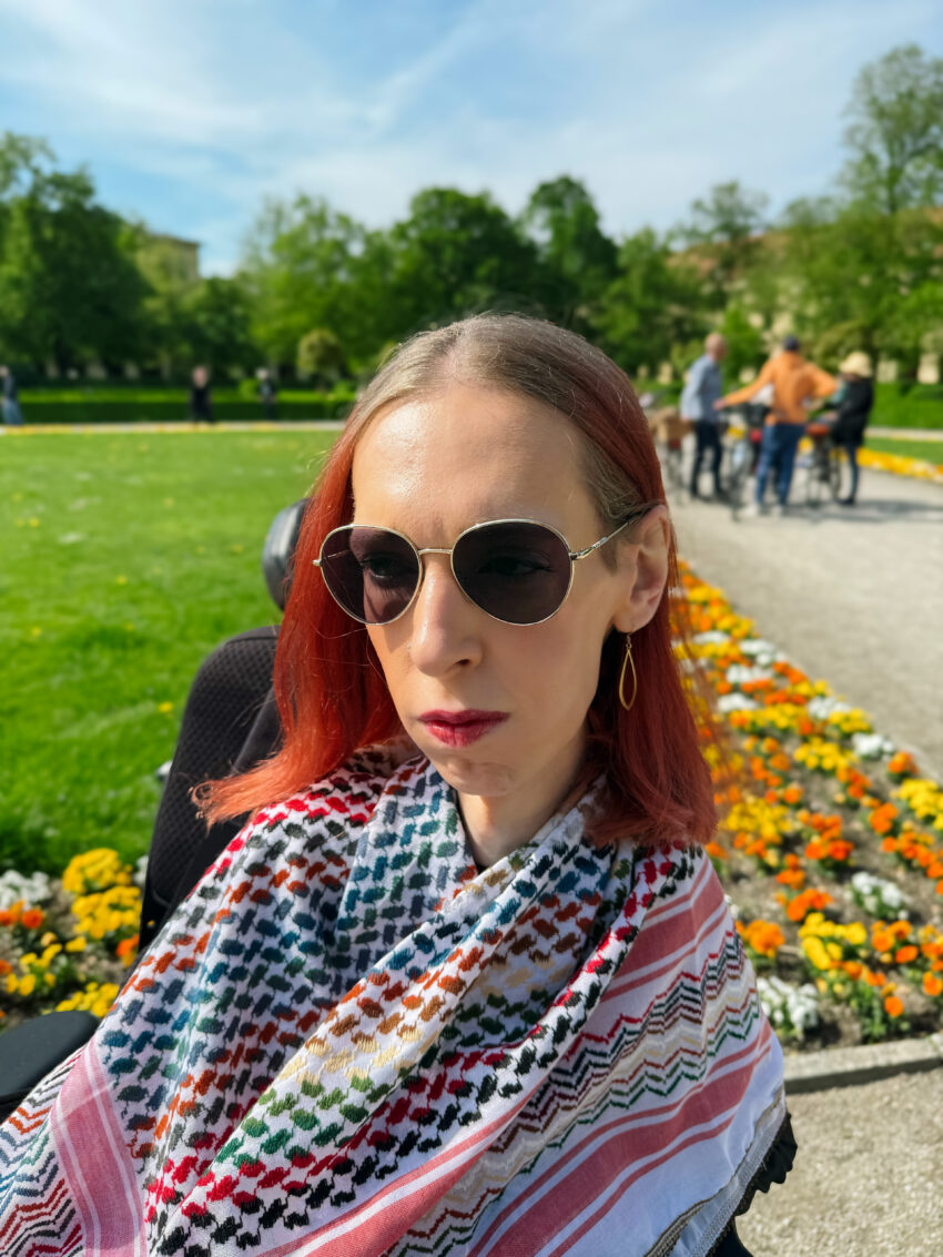 Ich stehe vor einem Blumenbeet, eingewickelt in eine bunte Kufiya. Mein Blick geht nachdenklich in die Ferne. Ich trage meine Haare offen und goldenen Schmuck. Mein Ausdruck ist äußerst nachdenklich und ernst.