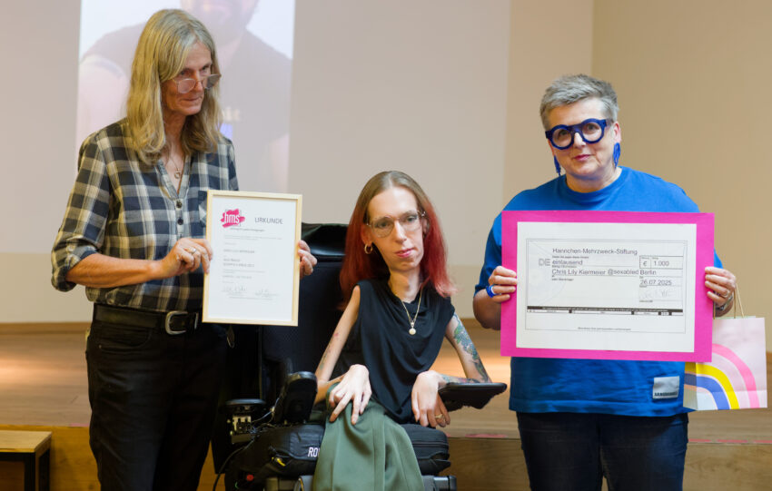 Ich sitze in Mitte eines Bühnenfotos zum Waltraud-Schiffels-Preis 2025. Links neben ihr steht Janka Kluge mit langen, grauen Haaren und karierter Bluse, die die Urkunde zeigt. Rechts neben mir steht Karen Noltevmit kurzen grauen Haaren, runder blauer Brille und blauem T-Shirt, die einen symbolischen Spendenscheck in pinkem Rahmen hochhält. Auf dem Scheck steht: „Hannchen-Mehrzweck-Stiftung – 1000 Euro für Chris Lily Kiermeier @sexabled“. Alle drei schauen in die Kamera. Im Hintergrund ist ein leicht verschwommenes Foto auf einer Leinwand zu erkennen.