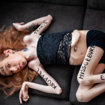 Die Fotografie zeigt mich mit langen, rötlich-blonden Haaren, wie ich seitlich auf einem dunklen Sofa liege. Ich trage ein schwarzes, bauchfreies Spitzentop und schwarze Shorts. Auf meiner Haut sind in großen, schwarzen Großbuchstaben verschiedene Beleidigungen geschrieben: „HURE“ auf der Stirn, „TRANSE“ auf dem rechten Unterarm, „KRÜPPEL“ auf dem linken Unterarm, „MISSGEBURT“ auf dem linken Oberschenkel und „EKLIGE“ auf dem linken Unterschenkel. Ich blicke ruhig, fast herausfordernd in Richtung Kamera. Das Bild wirkt roh und beklemmend und macht die Gewalt von Hate Speech visuell greifbar.