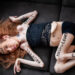 Die Fotografie zeigt mich mit langen, rötlich-blonden Haaren, wie ich seitlich auf einem dunklen Sofa liege. Ich trage ein schwarzes, bauchfreies Spitzentop und schwarze Shorts. Auf meiner Haut sind in großen, schwarzen Großbuchstaben verschiedene Beleidigungen geschrieben: „HURE“ auf der Stirn, „TRANSE“ auf dem rechten Unterarm, „KRÜPPEL“ auf dem linken Unterarm, „MISSGEBURT“ auf dem linken Oberschenkel und „EKLIGE“ auf dem linken Unterschenkel. Ich blicke ruhig, fast herausfordernd in Richtung Kamera. Das Bild wirkt roh und beklemmend und macht die Gewalt von Hate Speech visuell greifbar.