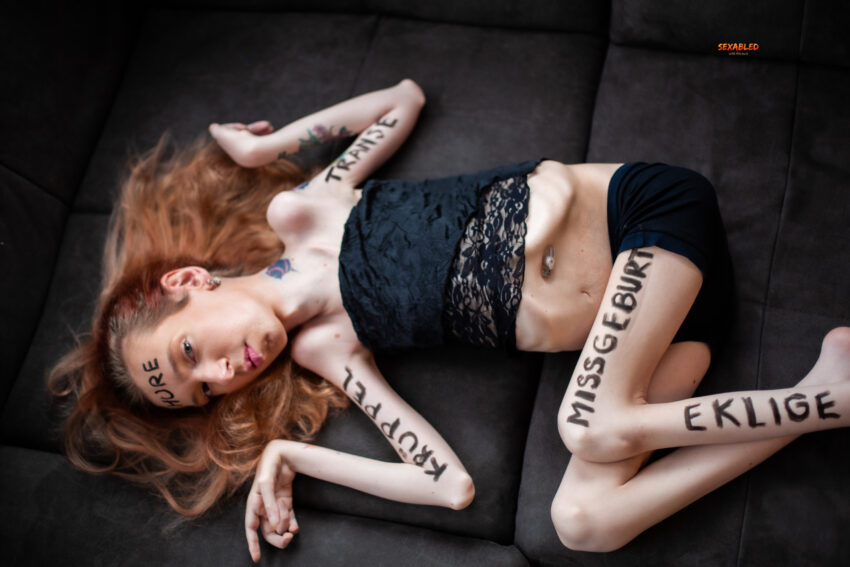 Die Fotografie zeigt mich mit langen, rötlich-blonden Haaren, wie ich seitlich auf einem dunklen Sofa liege. Ich trage ein schwarzes, bauchfreies Spitzentop und schwarze Shorts. Auf meiner Haut sind in großen, schwarzen Großbuchstaben verschiedene Beleidigungen geschrieben: „HURE“ auf der Stirn, „TRANSE“ auf dem rechten Unterarm, „KRÜPPEL“ auf dem linken Unterarm, „MISSGEBURT“ auf dem linken Oberschenkel und „EKLIGE“ auf dem linken Unterschenkel. Ich blicke ruhig, fast herausfordernd in Richtung Kamera. Das Bild wirkt roh und beklemmend und macht die Gewalt von Hate Speech visuell greifbar.